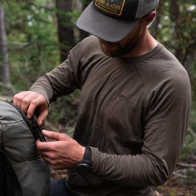 A backcountry hunter standing in the woods while wearing a Stone Glacier Chinook Merino Crew LS in tarmac and securing his backpack.