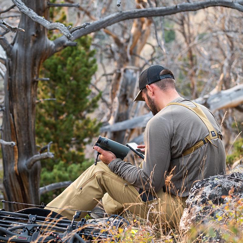 A backcountry hunter sitting on a ridge line and glassing with binoculars while wearing the Stone Glacier Chinook Merino Crew LS in tarmac.