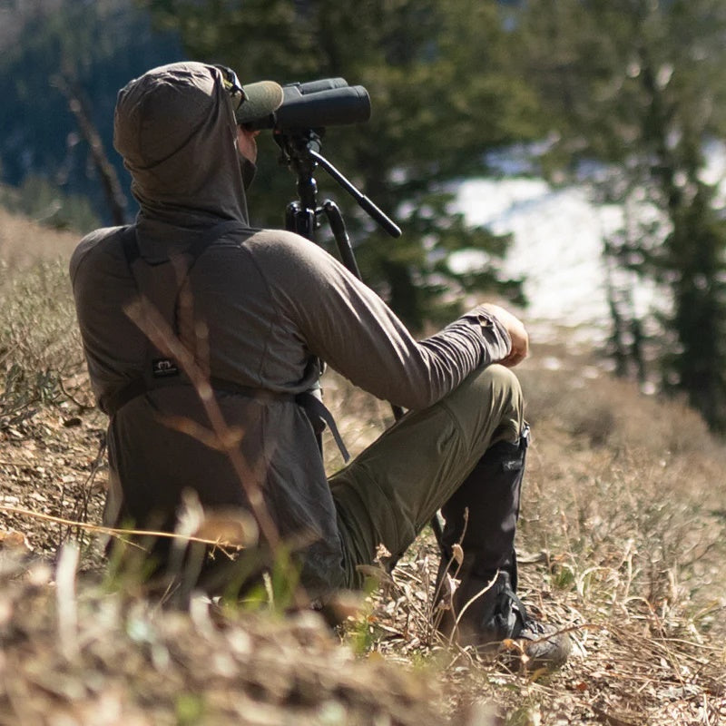 A backcountry hunter sitting on a mountain and glassing with his binoculars while wearing the Stone Glacier Chinook Merino Hoody.