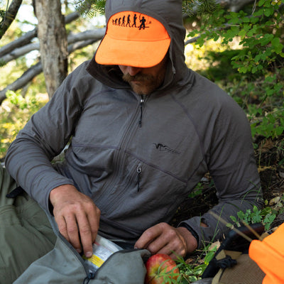 A backcountry hunter resting beneath a tree and sorting through his gear while wearing the Stone Glacier Chinook Merino Hoody.