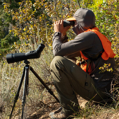 A backcountry hunter sitting in front of some brush while glassing through his binoculars and wearing the Stone Glacier Chinook Merino Hoody.