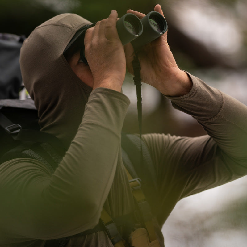 A backcountry hunter glassing upward through this binoculars amongst the trees while wearing the Stone Glacier Chinook Merino Hoody.