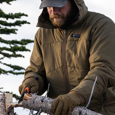 A man cutting down a tree with a handsaw while wearing the Stone Glacier Cirque Jacket in muskeg.