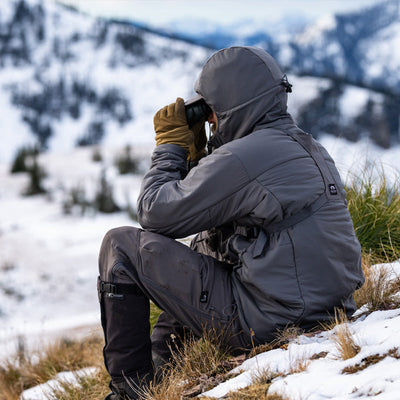 A backcountry hunter sitting on a hilltop amongst a snowy climate and glassing with binoculars while wearing the Stone Glacier Cirque Jacket in granite grey.