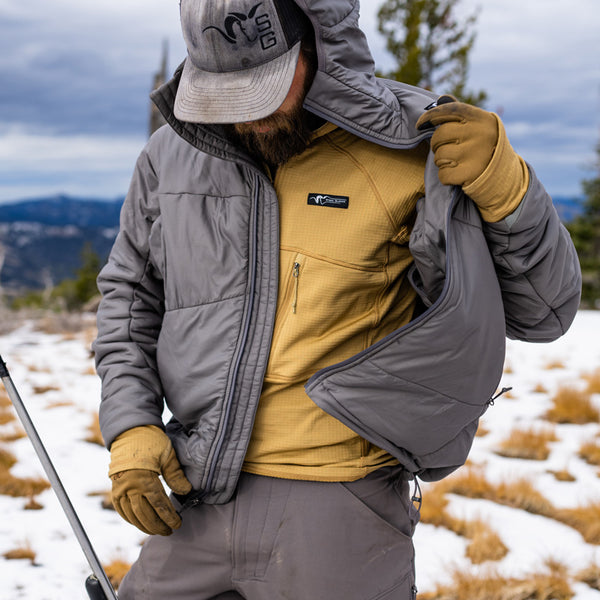 A backcountry hunter standing on top of a mountain while unzipping his Stone Glacier Cirque Jacket in granite grey and looking downward.