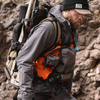 A backcountry rifle hunter walking along a cliff while wearing a backpack and the Stone Glacier De Havilland Jacket in granite grey.