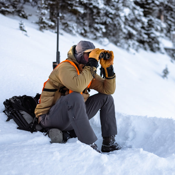 A backcountry hunter sitting on a snowy hillside and glassing with binoculars while wearing the Stone Glacier De Havilland Jacket in coyote.