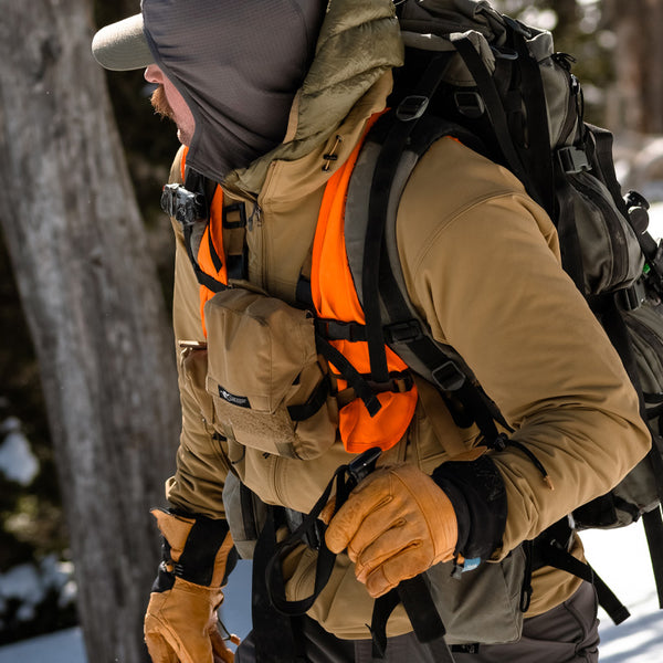 A backcountry hunter standing in the woods amongst a winter climate while wearing the Stone Glacier De Havilland Jacket in coyote.
