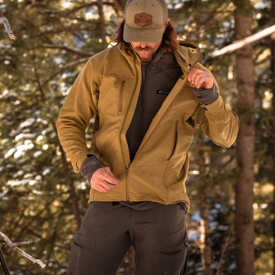 A backcountry hunter unzipping his Stone Glacier De Havilland Jacket in coyote while standing in front of an evergreen tree.