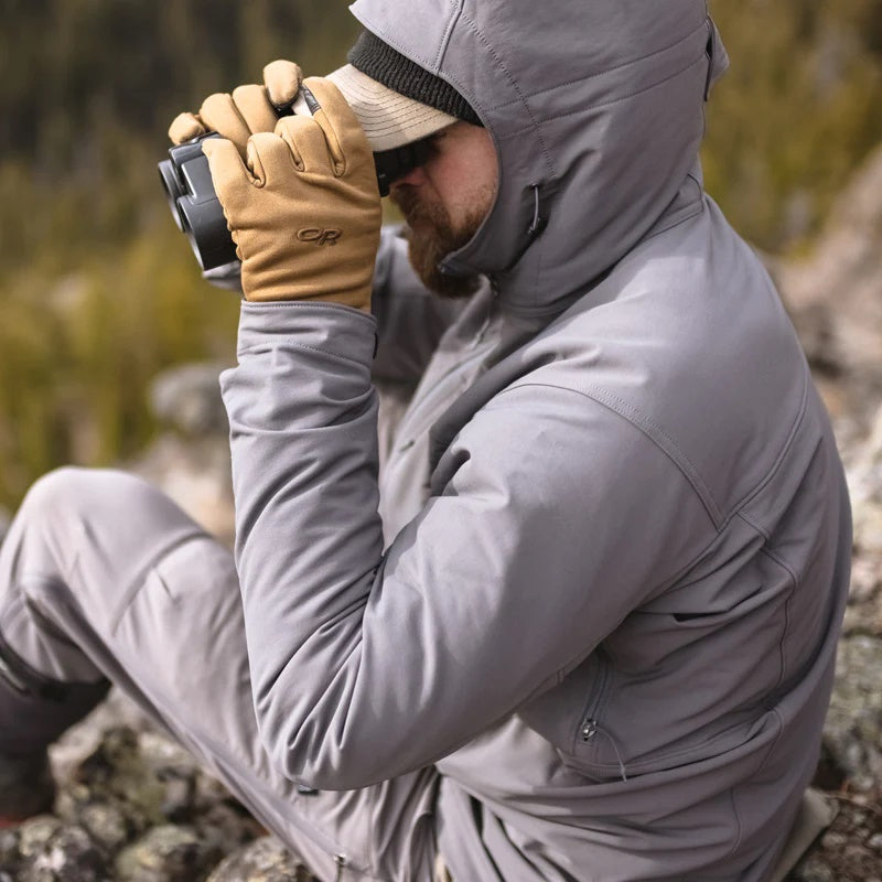 A backcountry hunter sitting on a rock while glassing with binoculars and wearing the Stone Glacier De Havilland Jacket in granite grey.