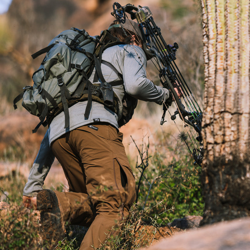 A backcountry archery hunter hiking uphill near saguaro cactus while wearing the Stone Glacier De Havilland LITE Pant in muskeg.
