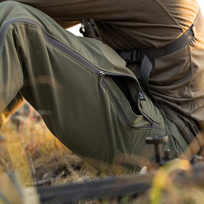 A close-up image of a backcountry hunter sitting on a remote hillside of grass while wearing the Stone Glacier De Havilland LITE Pants in tarmac.
