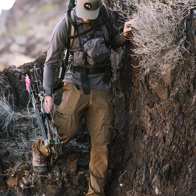 A backcountry archery hunter descending around a rock face with bow in hand while wearing the Stone Glacier De Havilland LITE Pants.