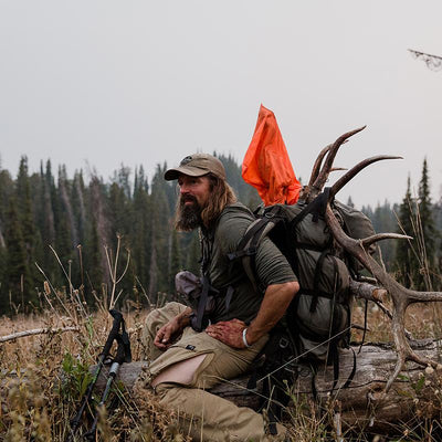 A backcountry hunter resting on a log while packing out a bull elk with his backpack and wearing a Stone Glacier DeHavilland Pant in granite grey.