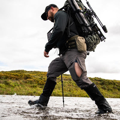 A backcountry hunter walking through a creek with his backpack on and wearing the Stone Glacier DeHavilland Pant in granite grey.
