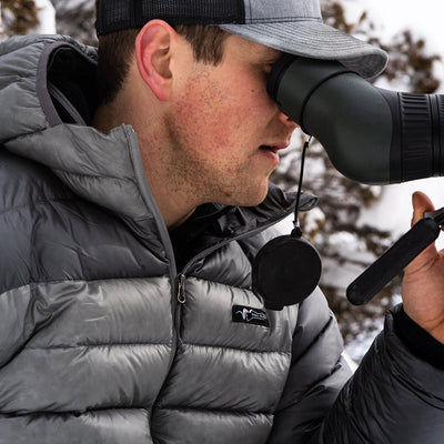 A close-up image of a backcountry hunter looking through a spotting scope while wearing a Stone Glacier Grumman Goose Down Jacket in granite grey.