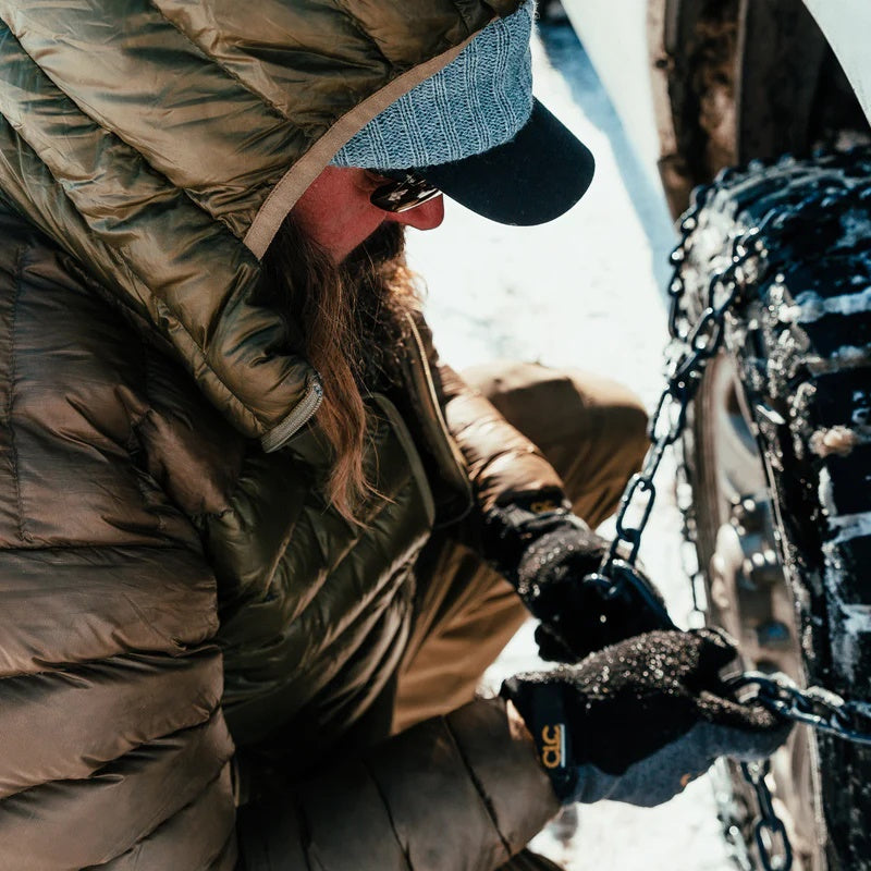 A backcountry hunter wearing the Stone Glacier Grumman Goose Down Jacket in coyote while installing snow chains on his pickup truck.