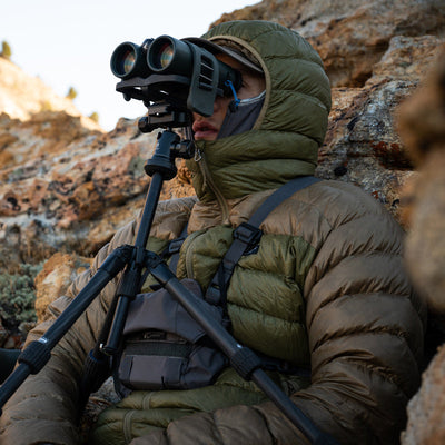 A backcountry hunter sitting in front of a rock cliff and glassing with binoculars while wearing a Stone Glacier Grumman Goose Down Jacket in coyote.