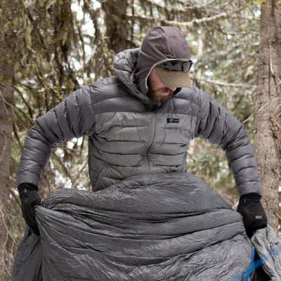 A backpacker laying out his sleeping bag in the woods while wearing a Stone Glacier Grumman Goose Down Jacket in granite grey.