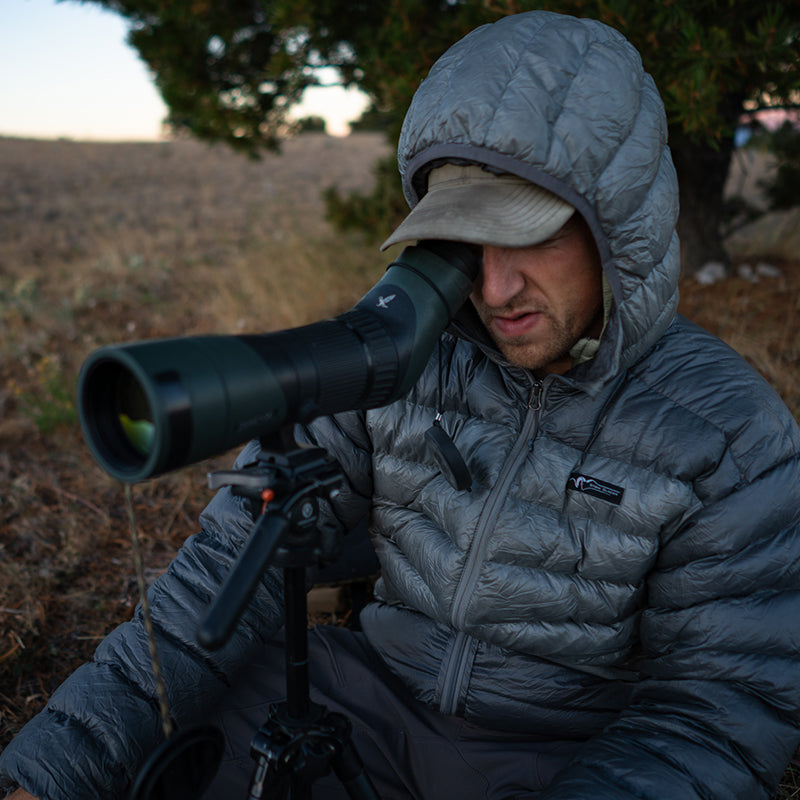 A backcountry hunter sitting on a ridge and glassing with a spotting scope on a tripod while wearing a Stone Glacier Grumman Goose Down Jacket in granite grey.