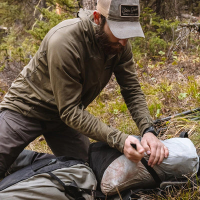 A backcountry hunter kneeling in a clearing and securing game bags to his backpack while wearing a Stone Glacier Helio Hoody.