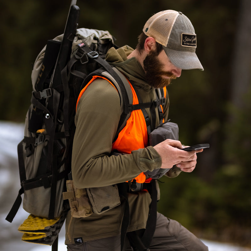 A backcountry rifle hunter navigating mountainous terrain with his smart phone while wearing the Stone Glacier Helio Hoody in fern.
