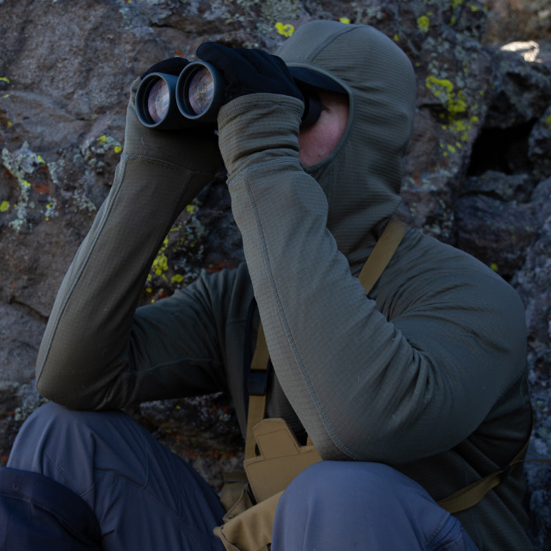 A backcountry hunter sitting near a boulder while glassing with binoculars and wearing a Stone Glacier Helio Hoody.