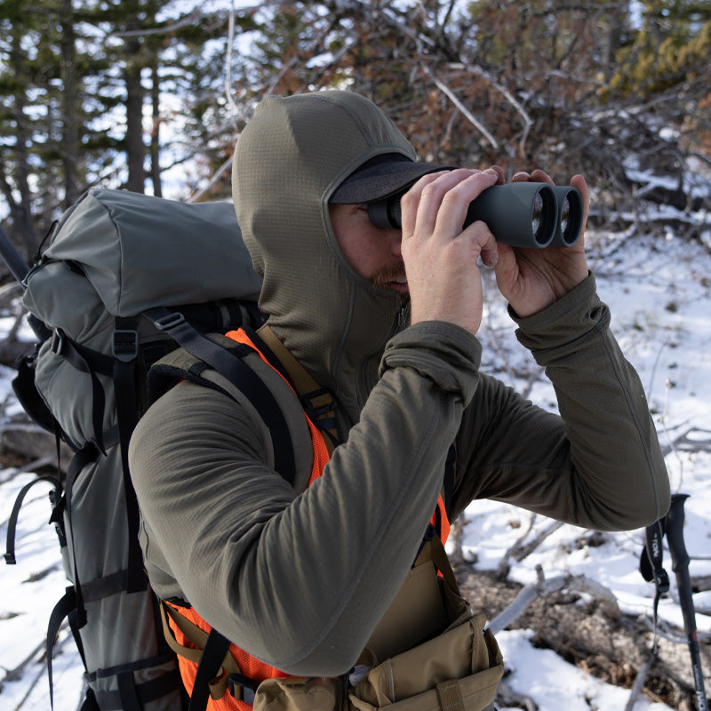 A backcountry hunter glassing with binoculars while standing in the snowy mountains and wearing the Stone Glacier Helio Hoody in fern.