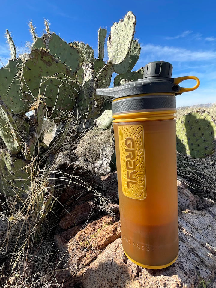 The GRAYL Geopress in coyote amber positioned above a rock and in front of cactus on a sunny day.