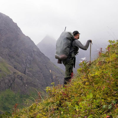 A backcountry hunter hiking on a steep mountainside in the high alpine while wearing the Stone Glacier Rain Cover over his backpack.