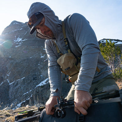 A backcountry hunter wearing a Stone Glacier Synthetic Hoody in stone grey while strapping compound bow to his backpack.