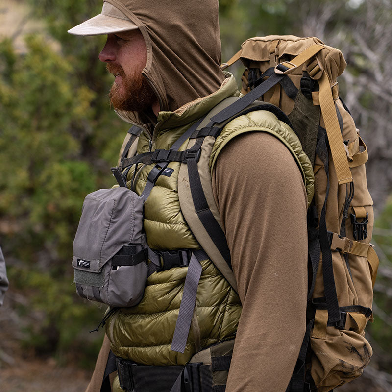 A backcountry hunter standing amongst the trees of a mountain landscape while wearing the Stone Glacier Synthetic Hoody in muskeg with backpack, vest, and binocular harness.