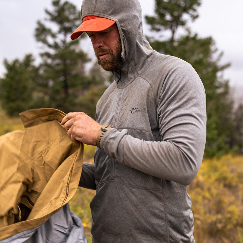 A backcountry hunter standing in a mountainous landscape while wearing the Stone Glacier Synthetic Hoody in stone grey and folding his rain gear.