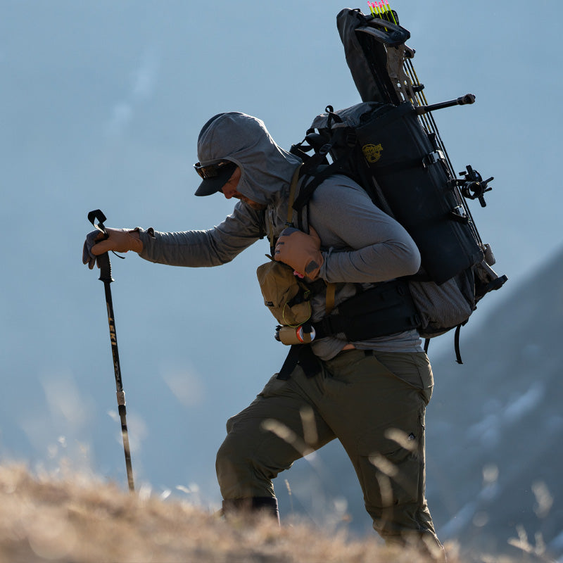 A backcountry hunter reaching the summit of a high mountain while wearing the Stone Glacier Synthetic Hoody in stone grey and heavy backpack.
