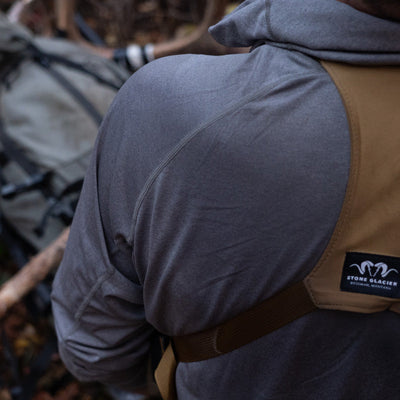 The rear left shoulder of a backcountry hunter who is wearing the Stone Glacier Synthetic Hoody in stone grey along with a binocular harness.