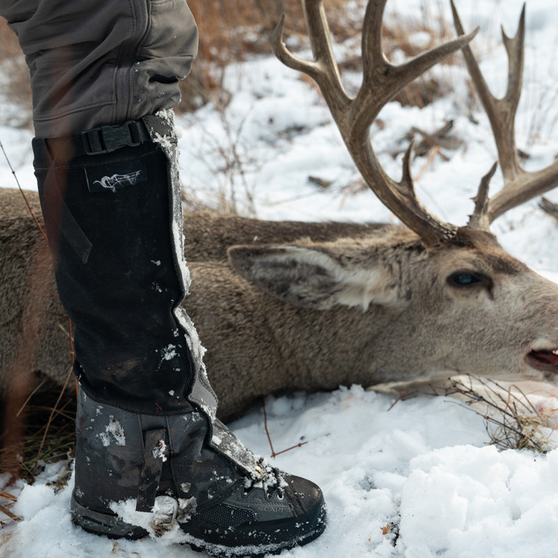 A close-up image of the Stone Glacier SQ2 Alpine Gaiters being worn by a hunter standing in front of his downed buck lying in the snow.