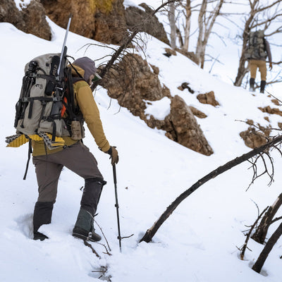 Two backcountry hunters hiking up a snowy hillside while wearing the Stone Glacier SQ2 Alpine Gaiters.