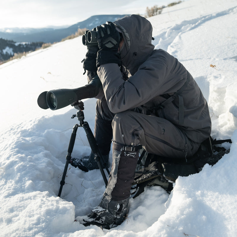 A backcountry hunter sitting in the snow while glassing through his binoculars and wearing the Stone Glacier SQ2 Alpine Gaiters.
