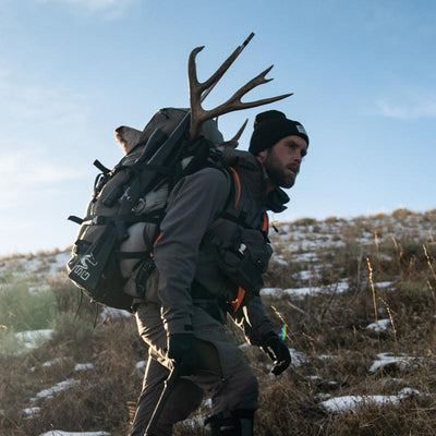 A backcountry hunter hiking the mountain while wearing the Stone Glacier Sky 5900 Backpack with a mule deer buck on top that he harvested.
