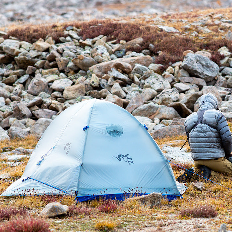 The Stone Glacier Sky Solus 1P Tent positioned in front a a rock scree on the side of a mountain in bright daylight with a backcountry hunter kneeling nearby.